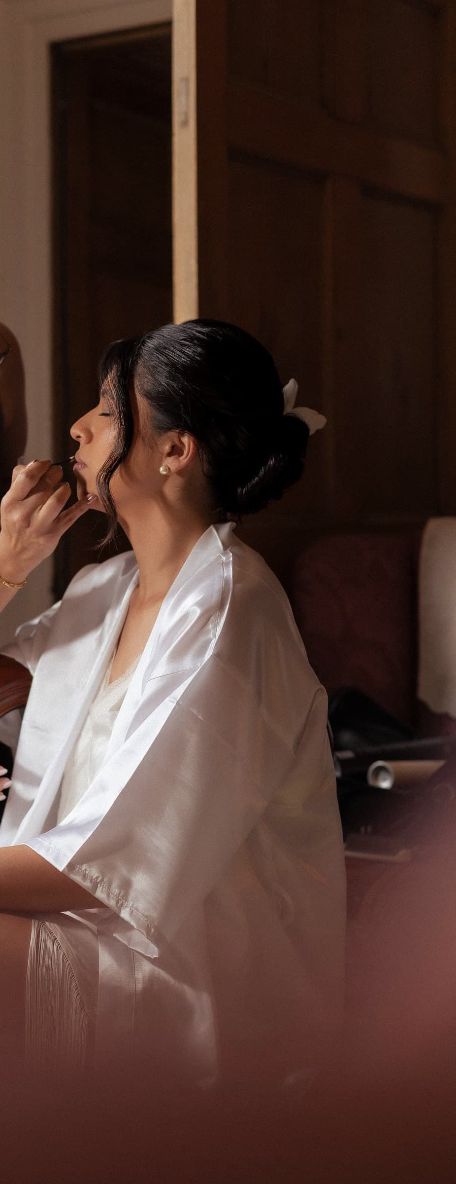 Bride in a white silk robe with her eyes closed while colour is applied to her lips.