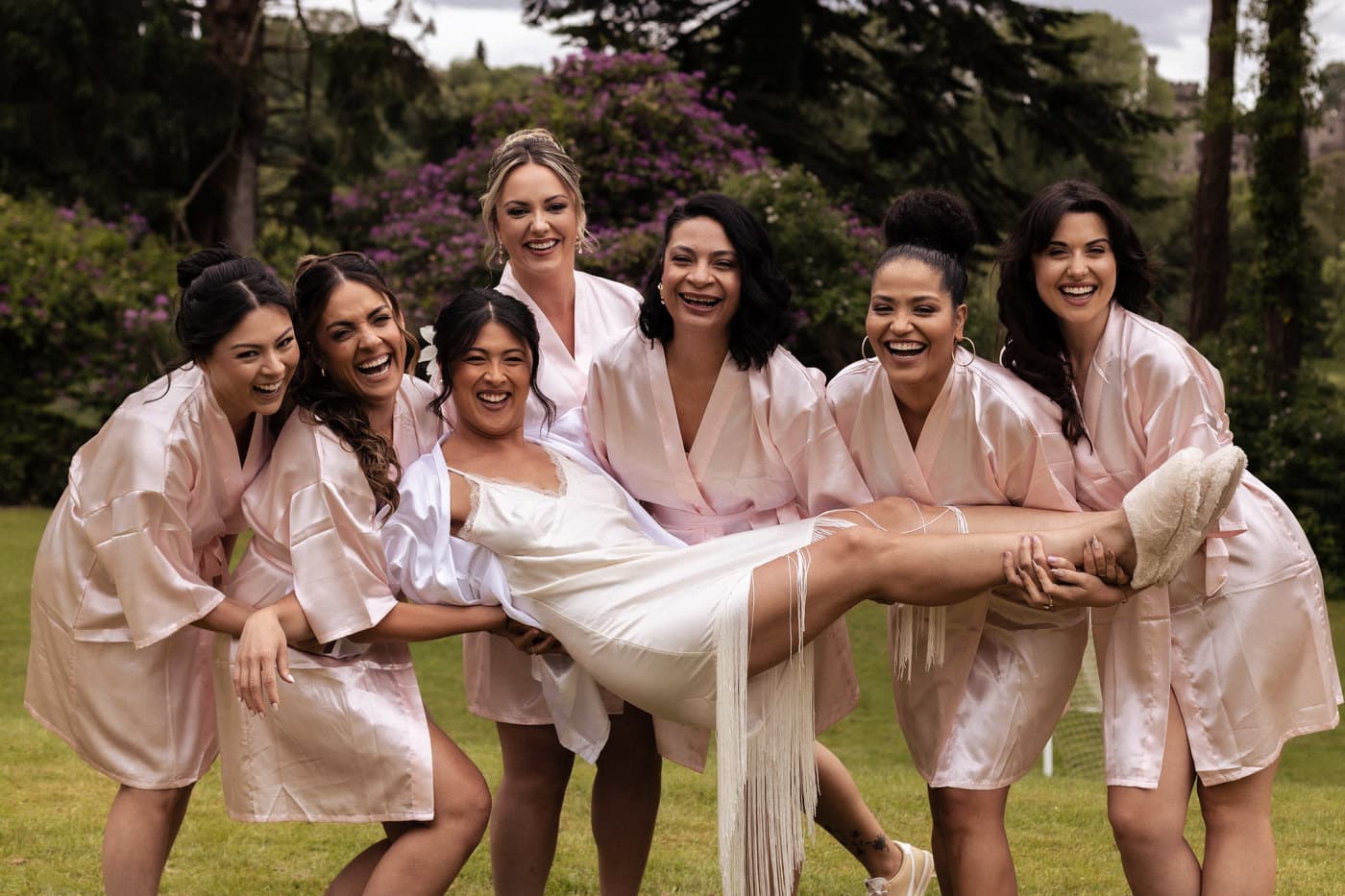 Bridesmaids in matching robes lifting the bride in a garden, all laughing.