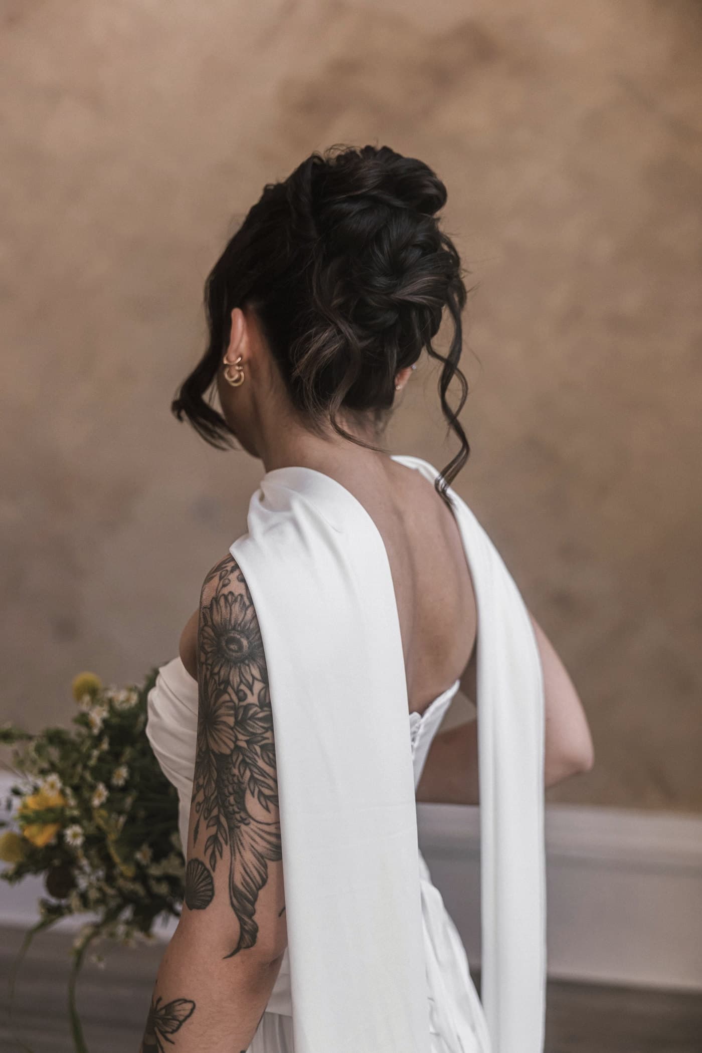 Bride seen from behind, hair in a low bun with small flowers, a tattoo across her shoulder.