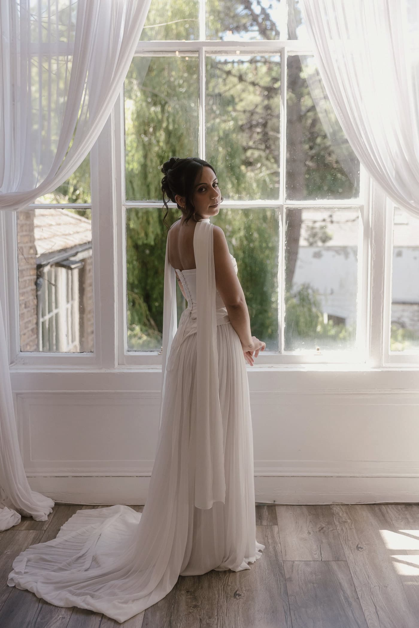 Bride standing at a window in a flowing dress, backlit by soft daylight.