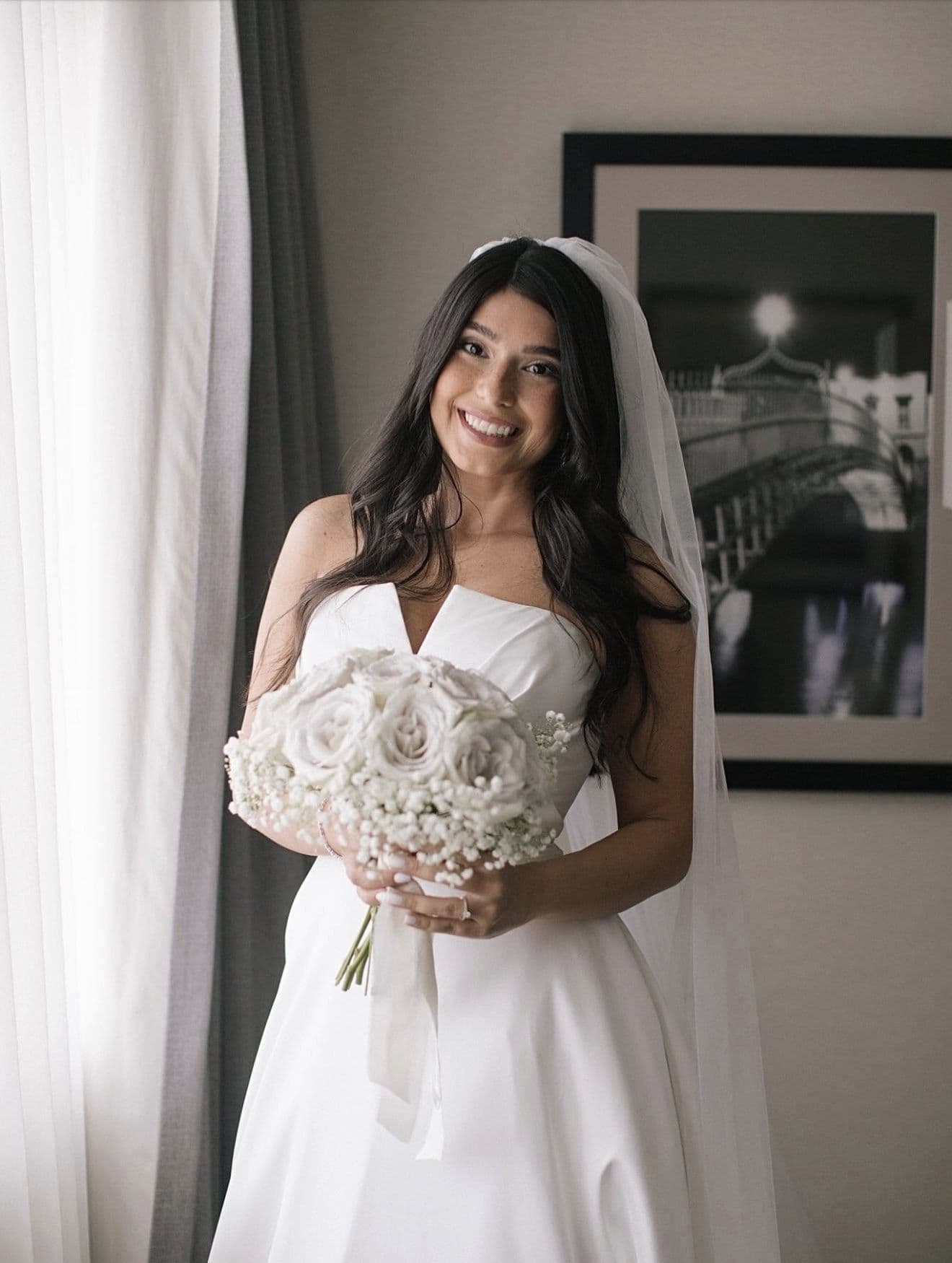 Smiling bride with soft waves and a veil holding a bouquet of white roses, standing in window light.