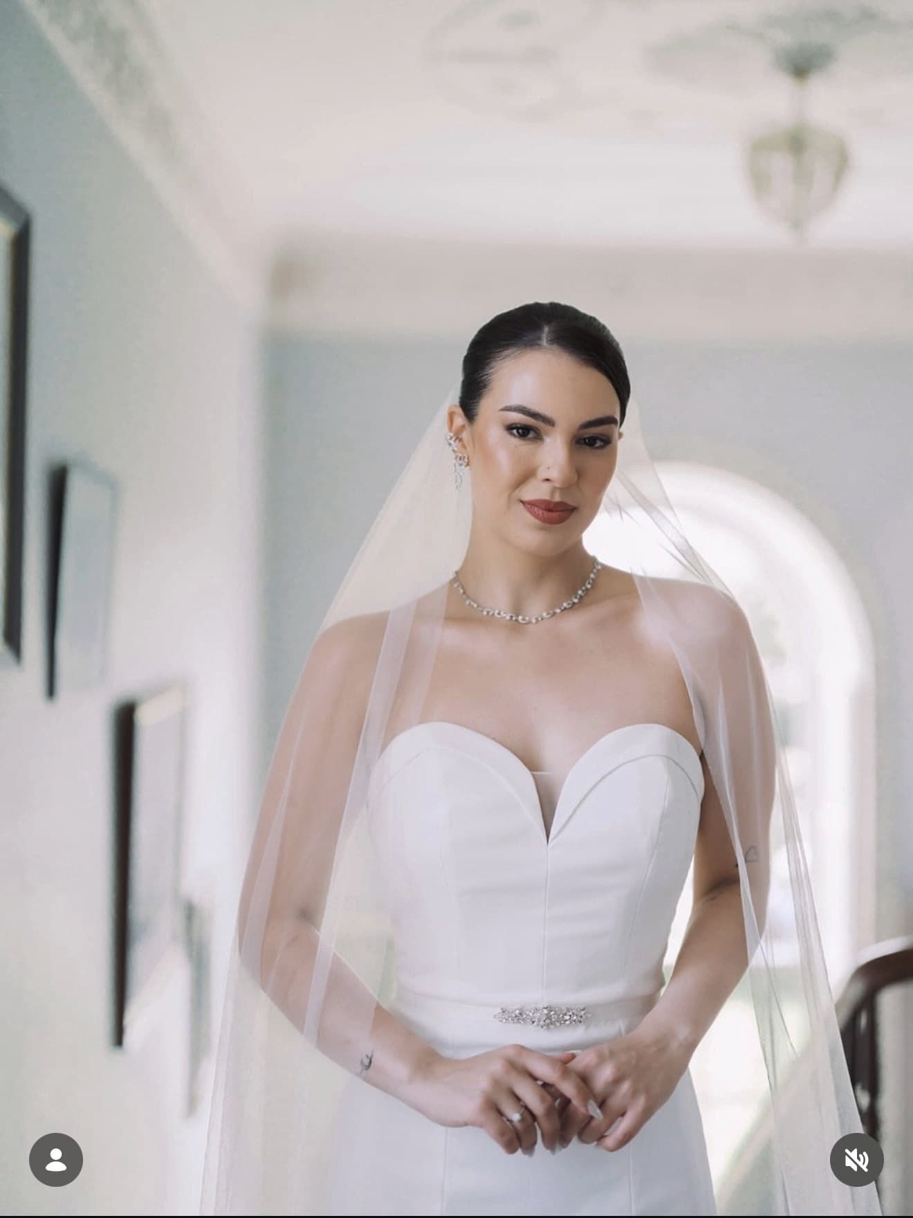 Bride walking a classical corridor under a chandelier, veil trailing behind her.